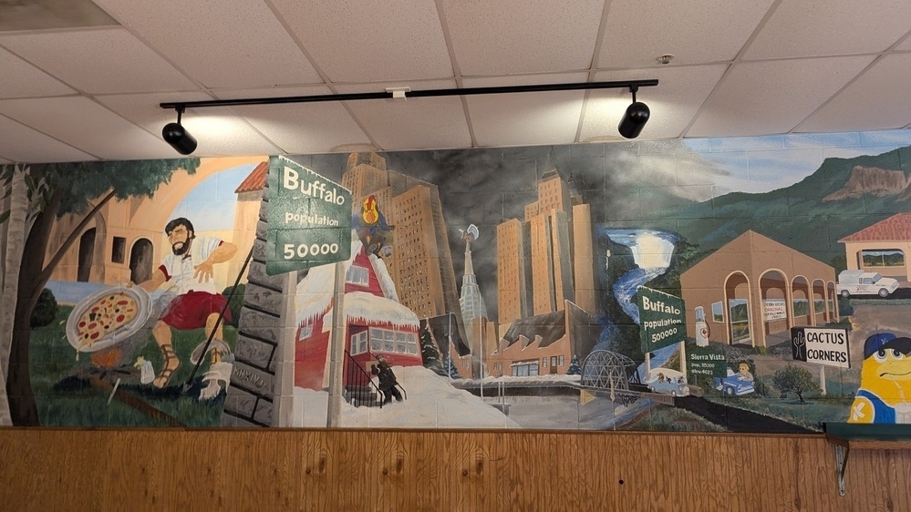 A colorful mural features various city landmarks, a person holding a pizza, and signs for Buffalo and Cactus Corners.