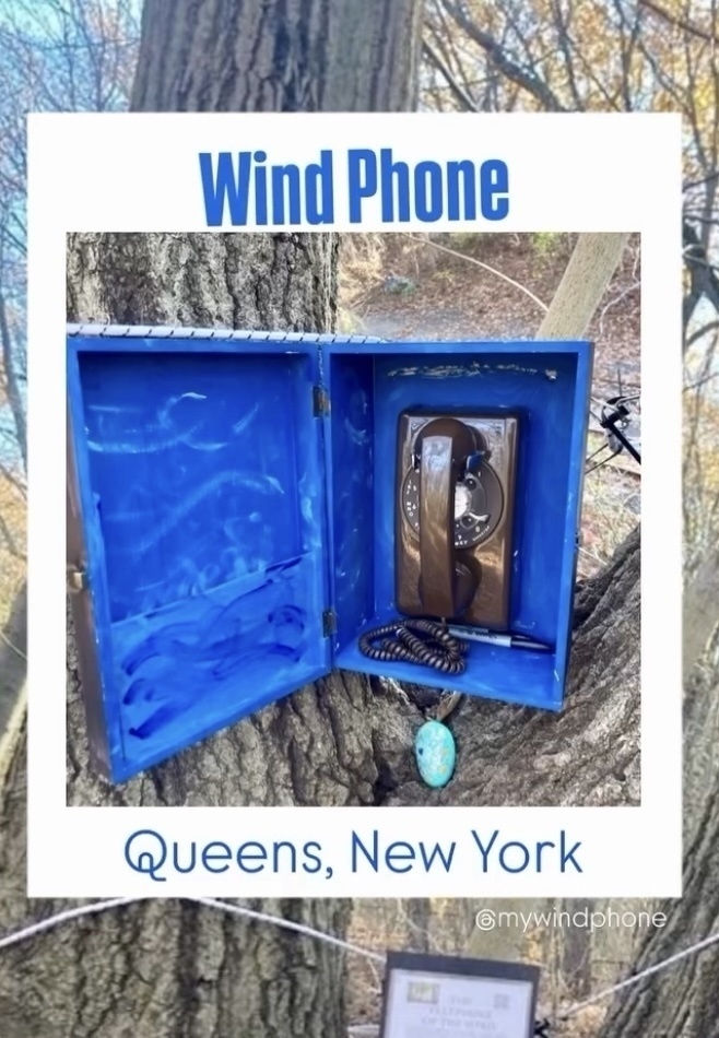 A vintage rotary phone is placed inside a blue box mounted on a tree, labeled Wind Phone, Queens, New York.