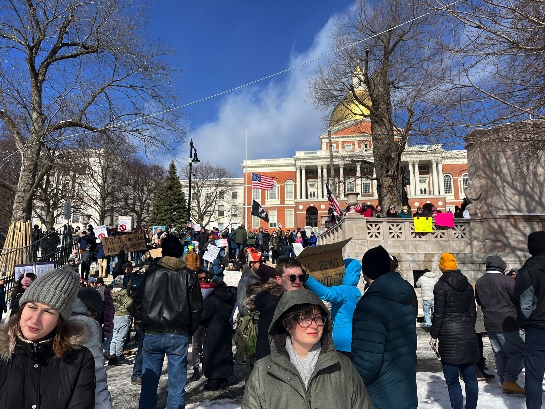 One protester has a sign saying ”first they came for the immigrants”. One has a sign saying ”Hey Elon, no coup for you”. One has a sign saying ”wake up and smell the fascism”. There are several American flags, a 1776 flag, and a POW flag.The Massachusetts State House is in the background. 