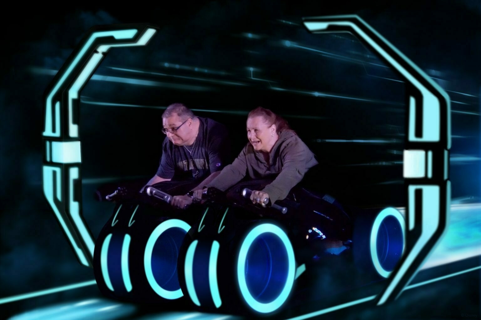 Two people are riding a futuristic motorcycle simulator with glowing blue neon lights.