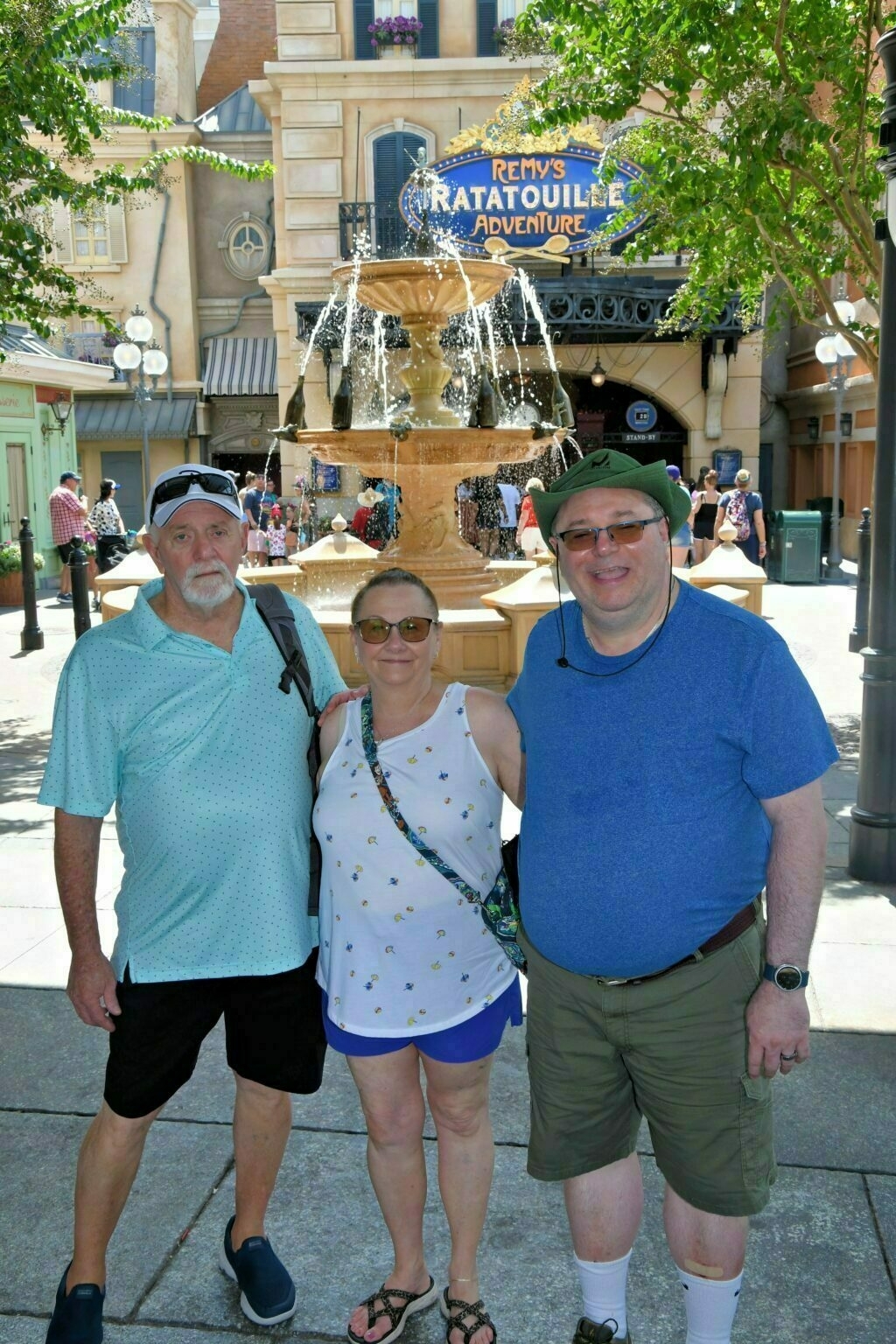 Three people are posing in front of a fountain at an amusement park, with a sign for Remy's Ratatouille Adventure in the background.