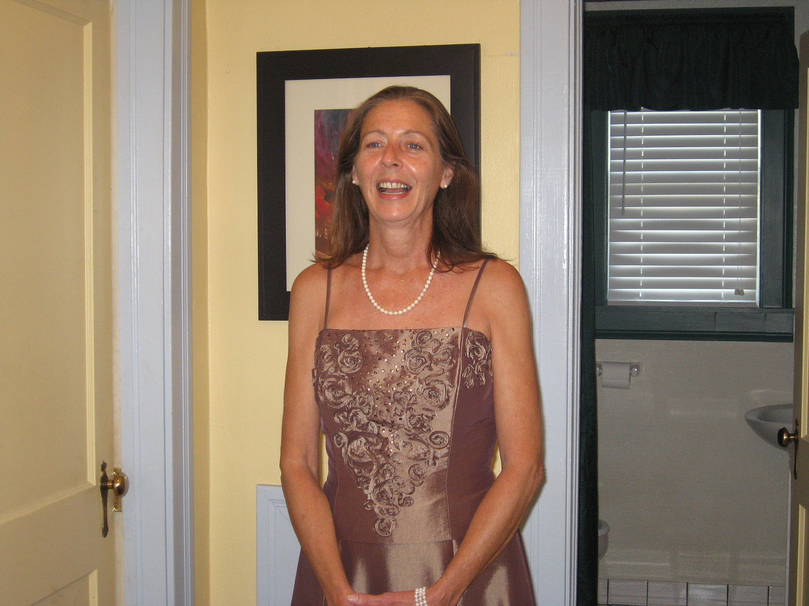 A woman in a brown dress is standing and smiling indoors near a bathroom.