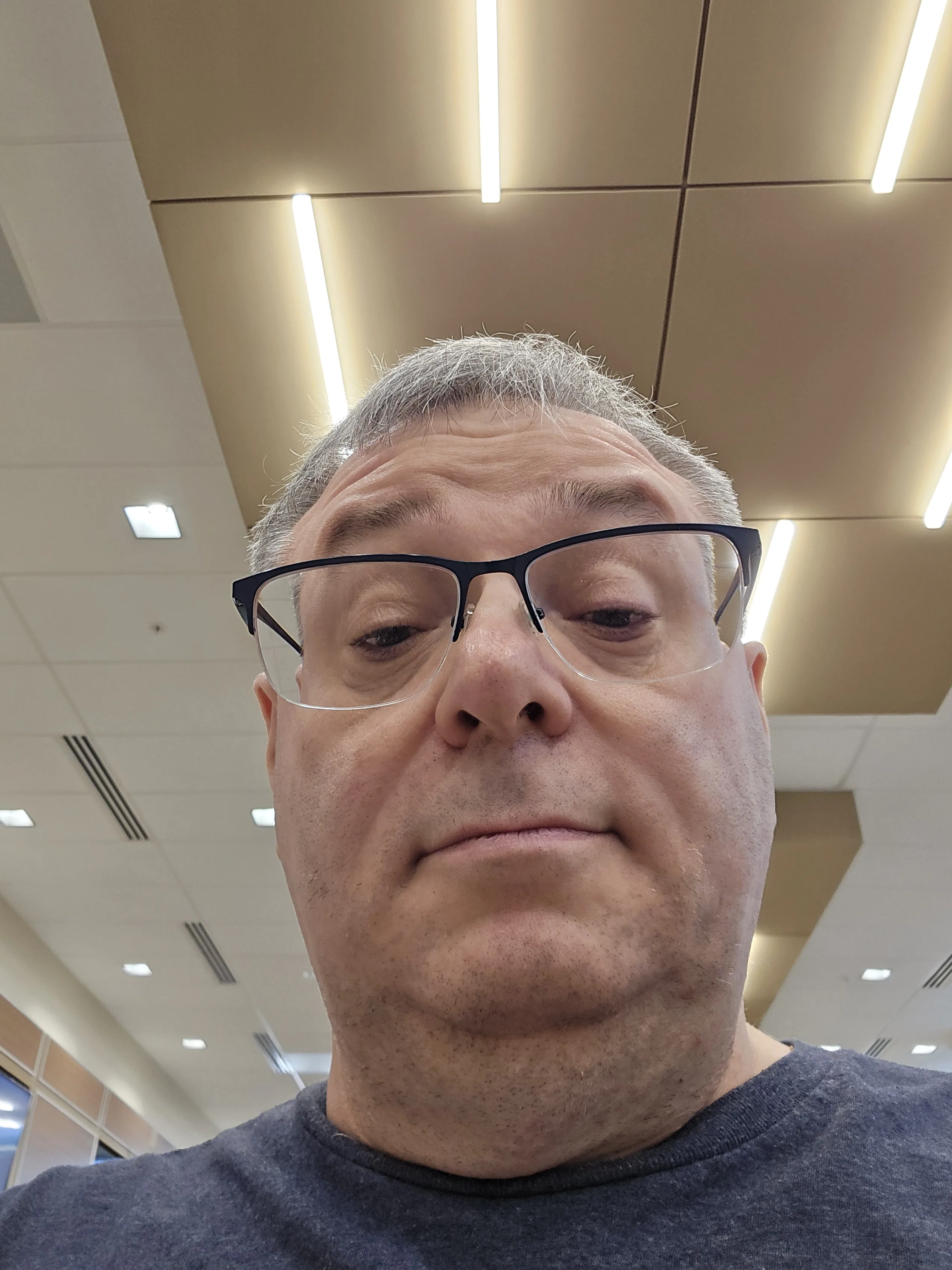 Close-up selfie of a middle-aged man with short gray hair and light skin, wearing rectangular black-framed glasses. The photo is taken from a low angle, capturing the lower half of his face prominently, including his nose, mouth, and chin, with a neutral expression. He is wearing a dark blue shirt. The background shows an indoor setting with a ceiling featuring beige panels and long, bright fluorescent lights arranged in parallel lines. Additional ceiling elements include white square tiles and air vents. The lighting is bright and evenly distributed.