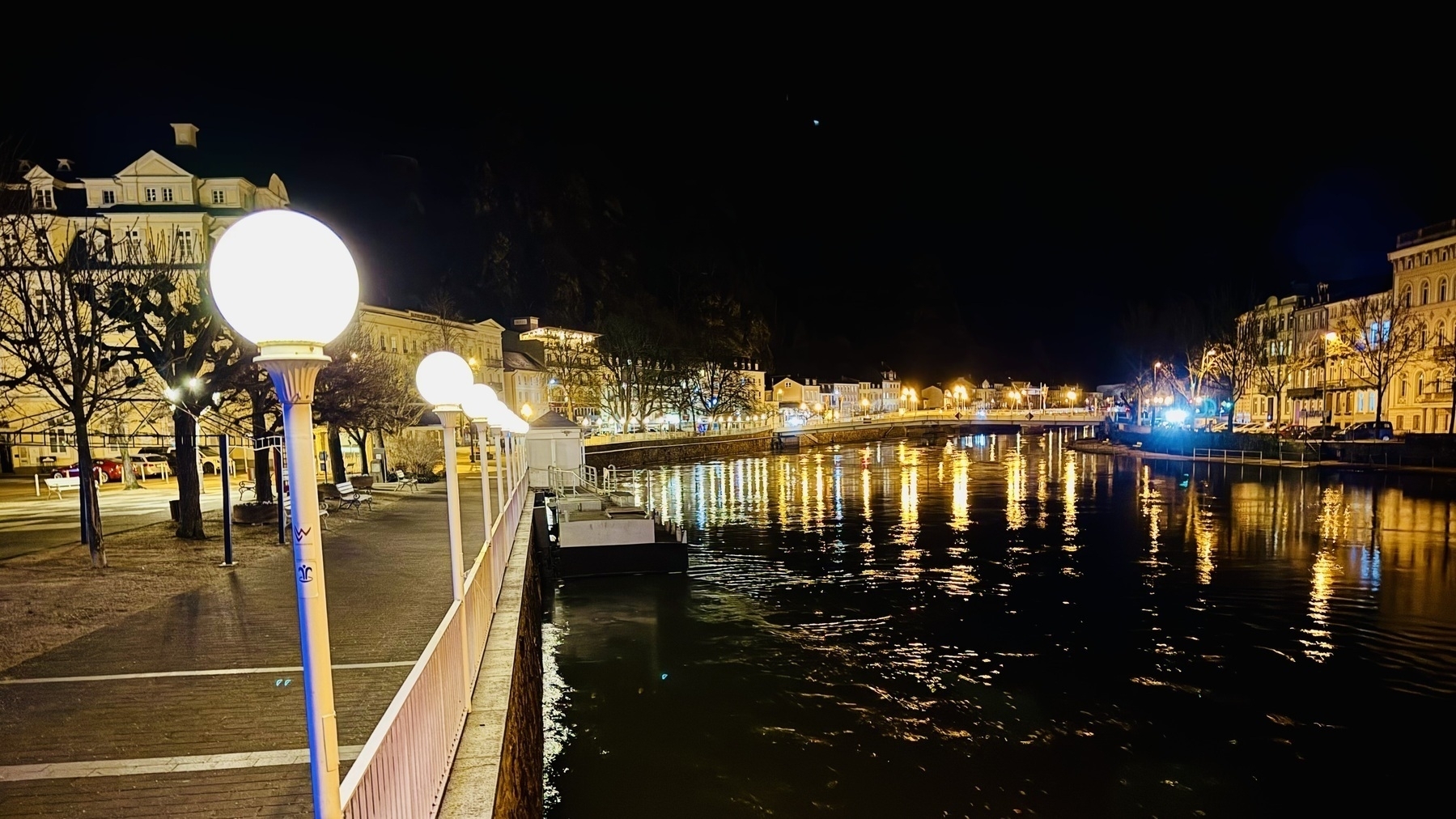 This is a nighttime photograph taken in Bad Ems, showcasing a scenic riverside view. The brightly illuminated street lamps along the promenade cast a warm glow over the area, with their light reflecting beautifully on the calm surface of the river. Historical buildings with intricate architectural details line the street on the left side, adding charm to the scene. The overall ambiance is serene and inviting, with the reflections on the water enhancing the tranquil atmosphere.