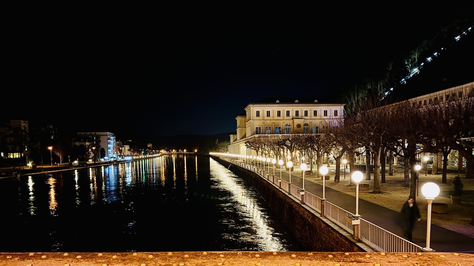 This is another stunning nighttime photograph from Bad Ems, focusing on a riverside promenade with beautifully aligned glowing street lamps. The river reflects the warm light, creating a picturesque golden shimmer on the water. On the right, a grand historical building stands illuminated, emphasizing its elegant architecture. Bare trees line the walkway, adding a sense of symmetry and charm to the scene. In the distance, the hill on the far right is faintly lit, enhancing the contrast between the natural and urban elements. The overall atmosphere is peaceful and captivating, showcasing the timeless beauty of Bad Ems.