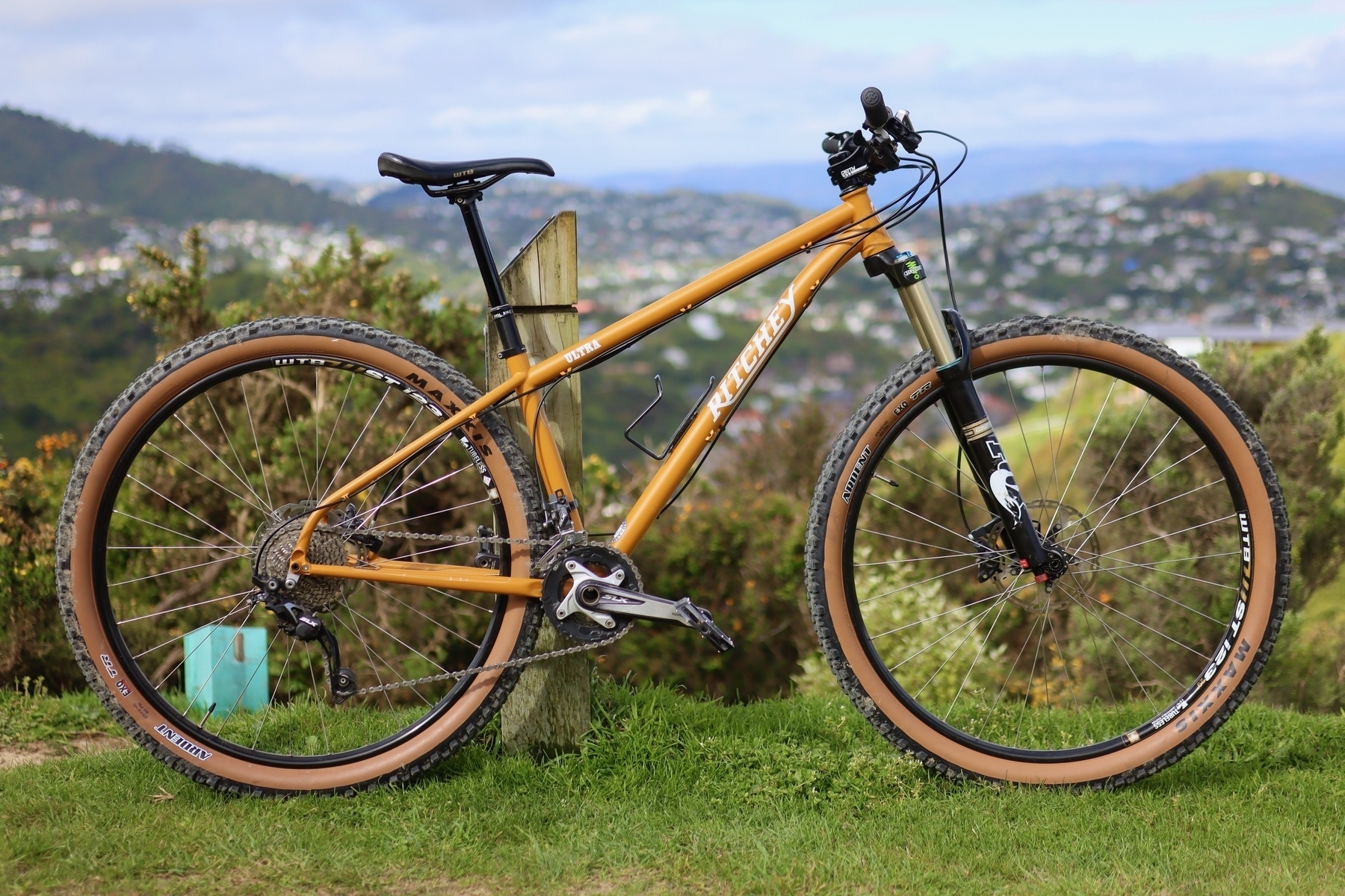 An orange Ritchey Ultra mountainbike is displayed side on, leaning on a track marker. In the background is some vegetation and in the distance some out of focus suburbs.