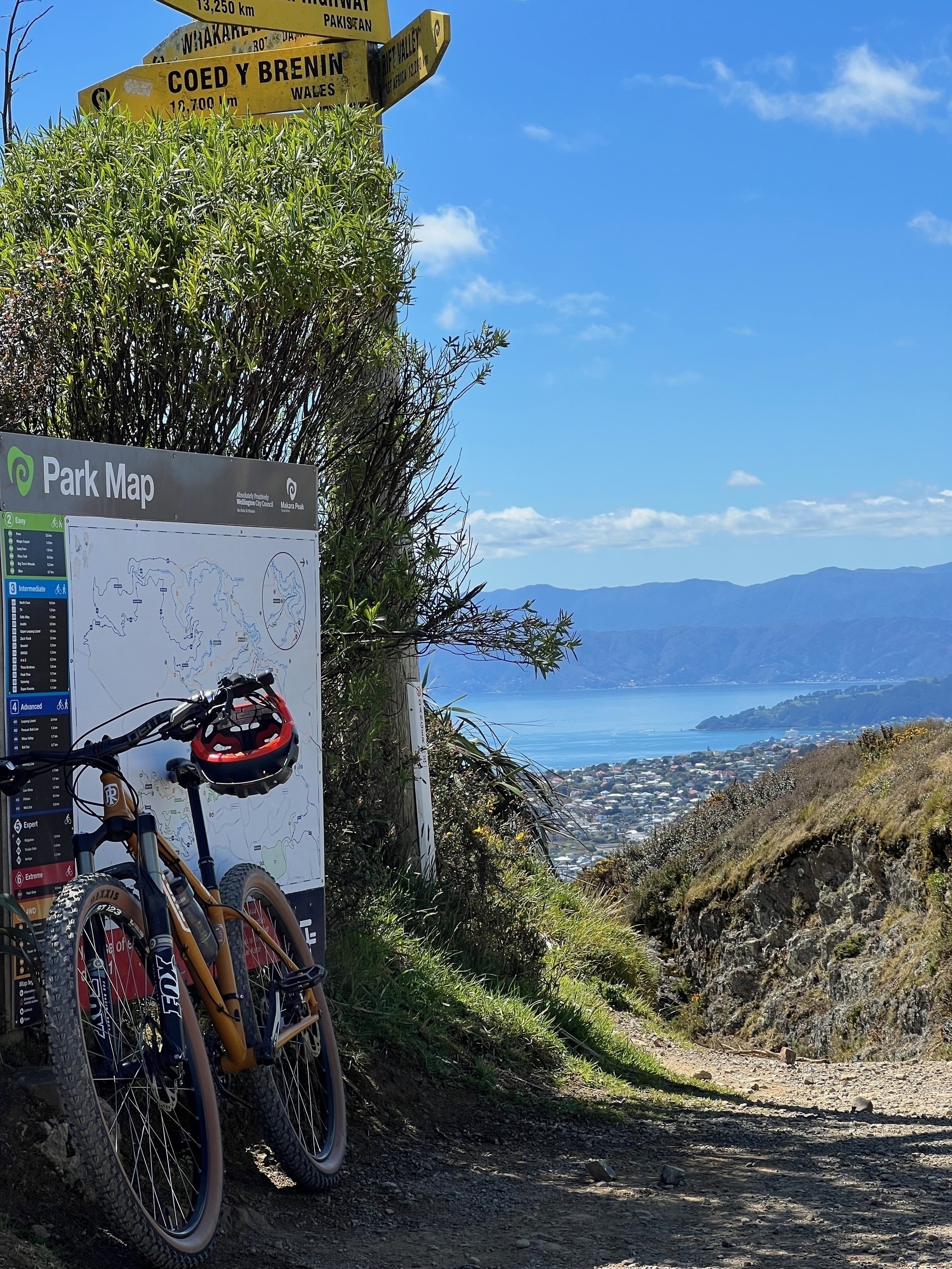 An orange bike is parked by a map of Makara Peak mountain bike park. Behind is the suburb of Karori, and in the distance, Wellington Harbour.
