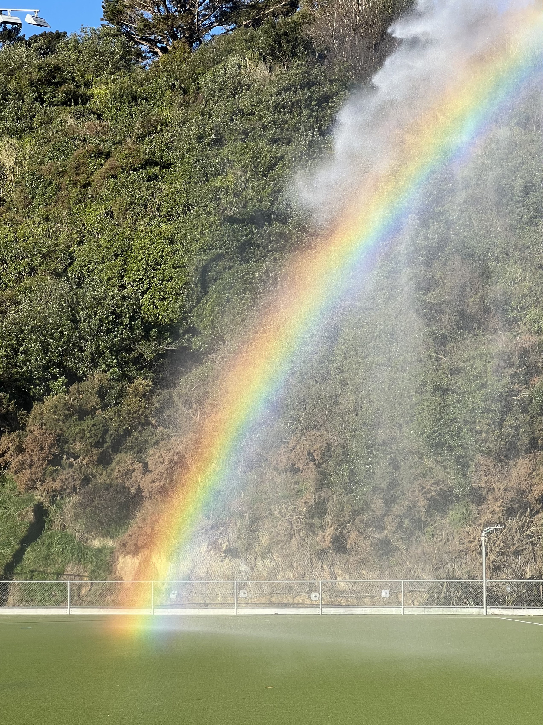 A vivid rainbow caused by Kate afternoon sunshine on a plume of water falling into an artificial grass field hockey pitch. In the background is a very steep hill covered in scrubby green vegetation, and some blue sky beyond is visible.&10;