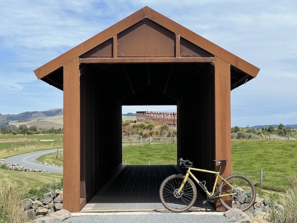 An american style covered bridge - but made of steel. In the distance, through the bridge, another bridge is visible. There is a yellow bike positiond in front of the closest bridge.