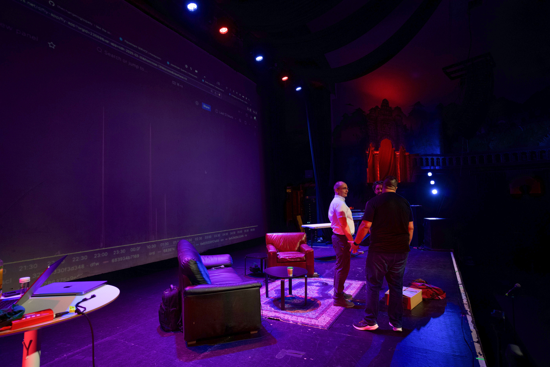 Auto-generated description: Two people are standing on a dimly lit stage with chairs, a table, and a rug, surrounded by theatrical lighting and shadows.