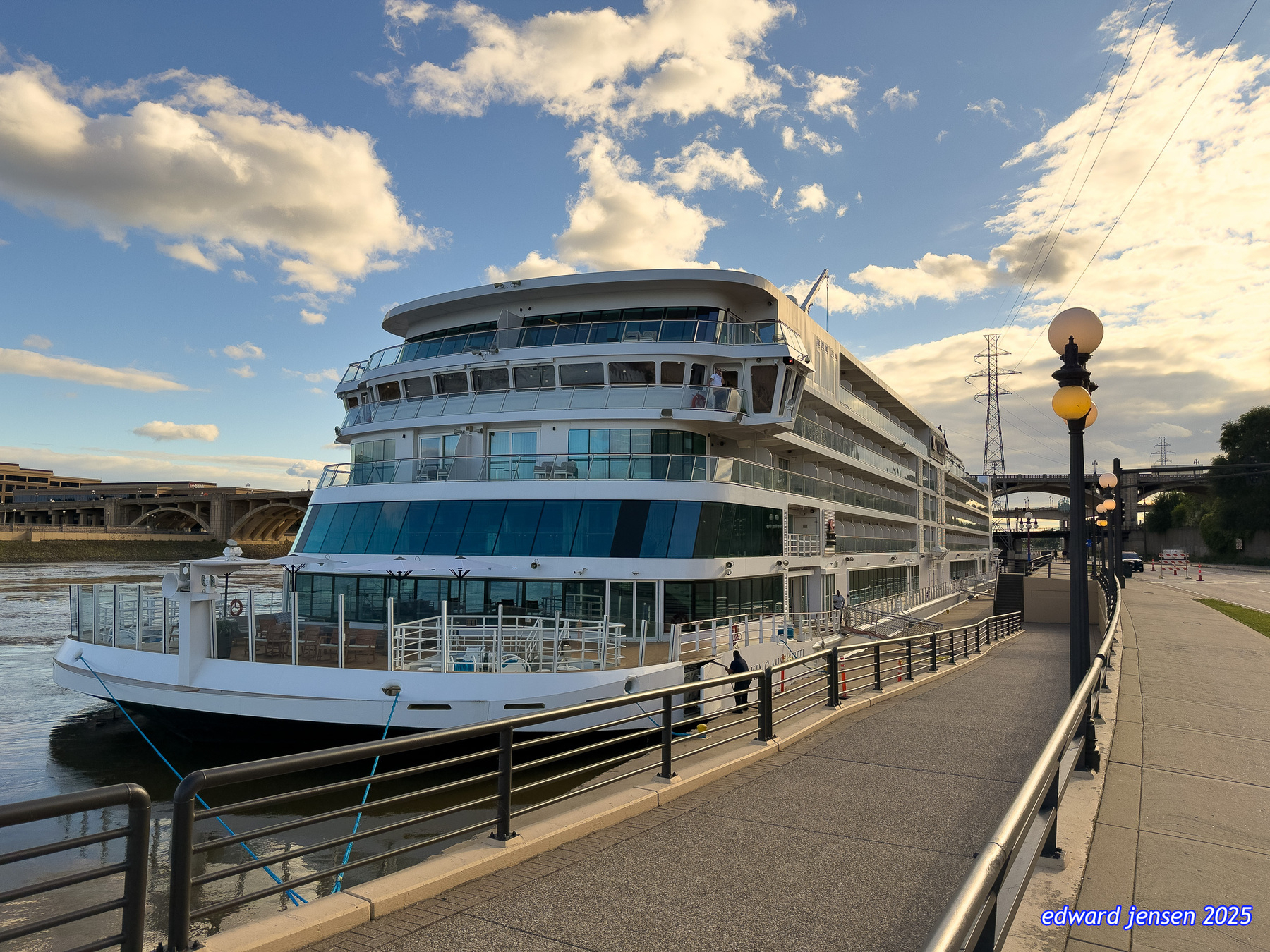 A large modern white cruise ship or river boat docked at a waterfront promenade with multiple decks, large windows, and curved architecture, moored alongside a paved walkway with metal railings and ornate lampposts under a dramatic cloudy sky with bridges visible in the background.