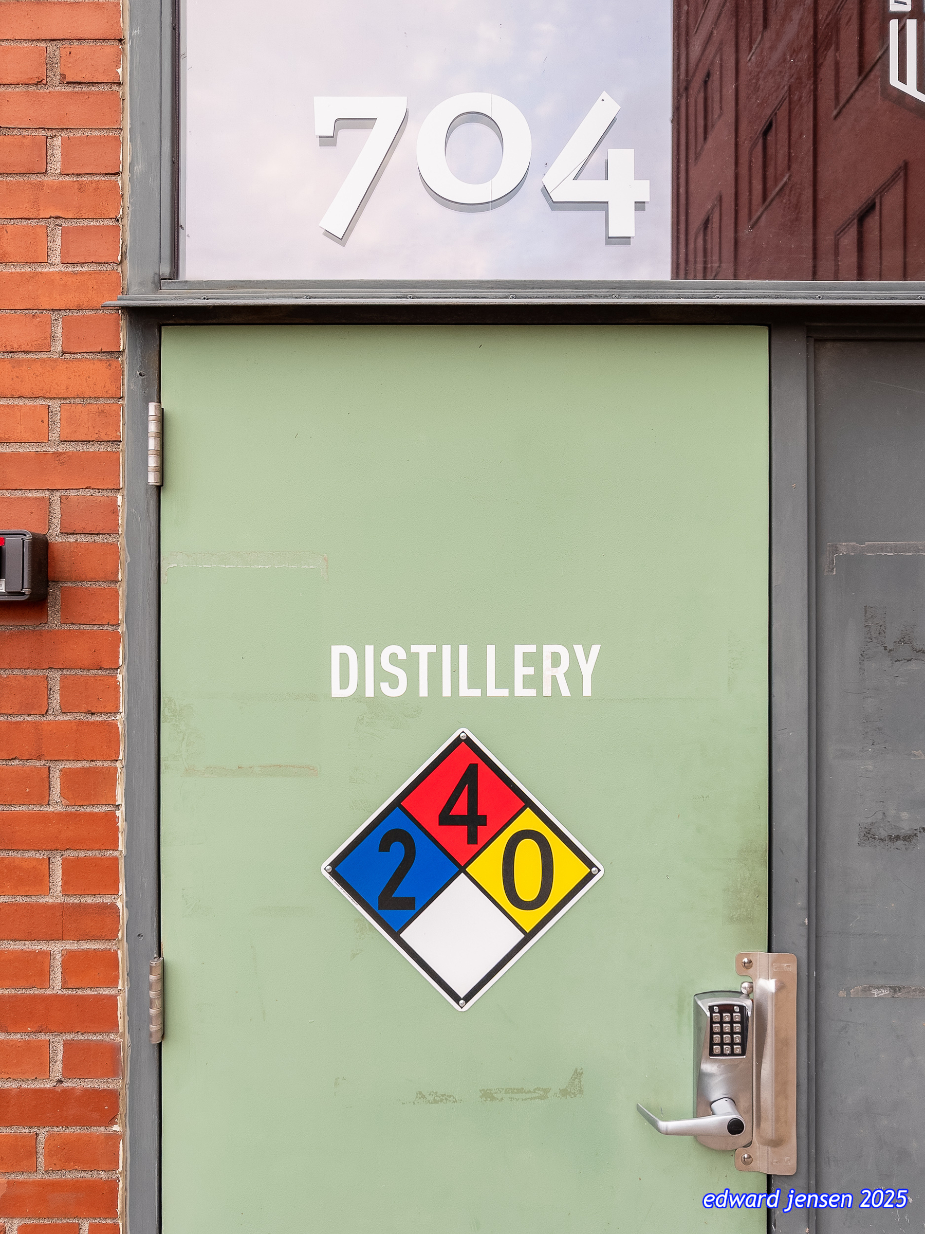 Photograph of a pale green door with the number 704 above it and an NFPA 704 hazard diamond sign reading \"DISTILLERY\" with ratings of 2 for health (blue), 4 for flammability (red), and 0 for reactivity (yellow), next to orange brick wall with electronic keypad lock visible.