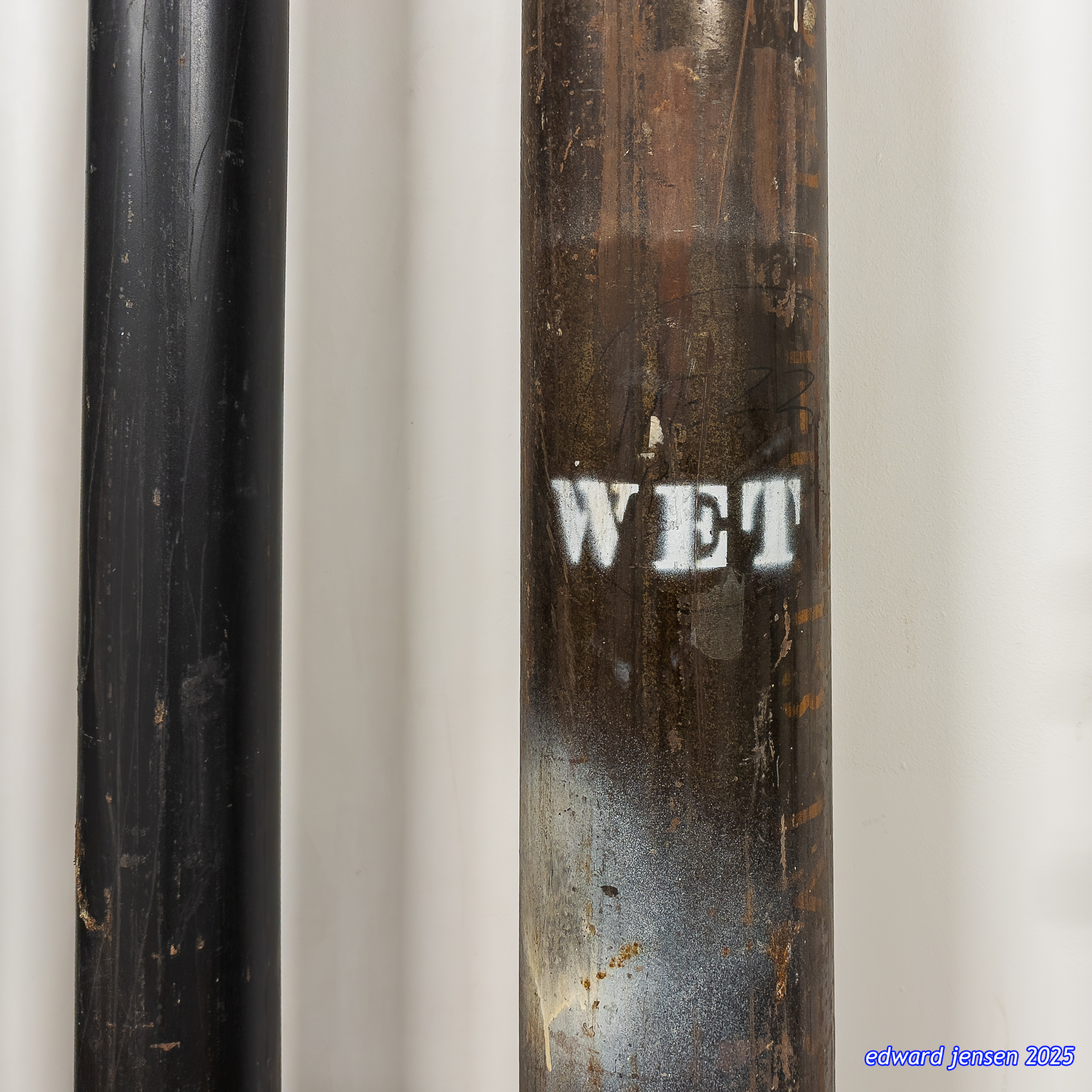 Close-up photograph of two vertical metal pipes against a light wall; the left pipe is black with scratches, the right pipe is weathered and rusted with the word WET stenciled in white paint. Watermark reads edward jensen 2025.