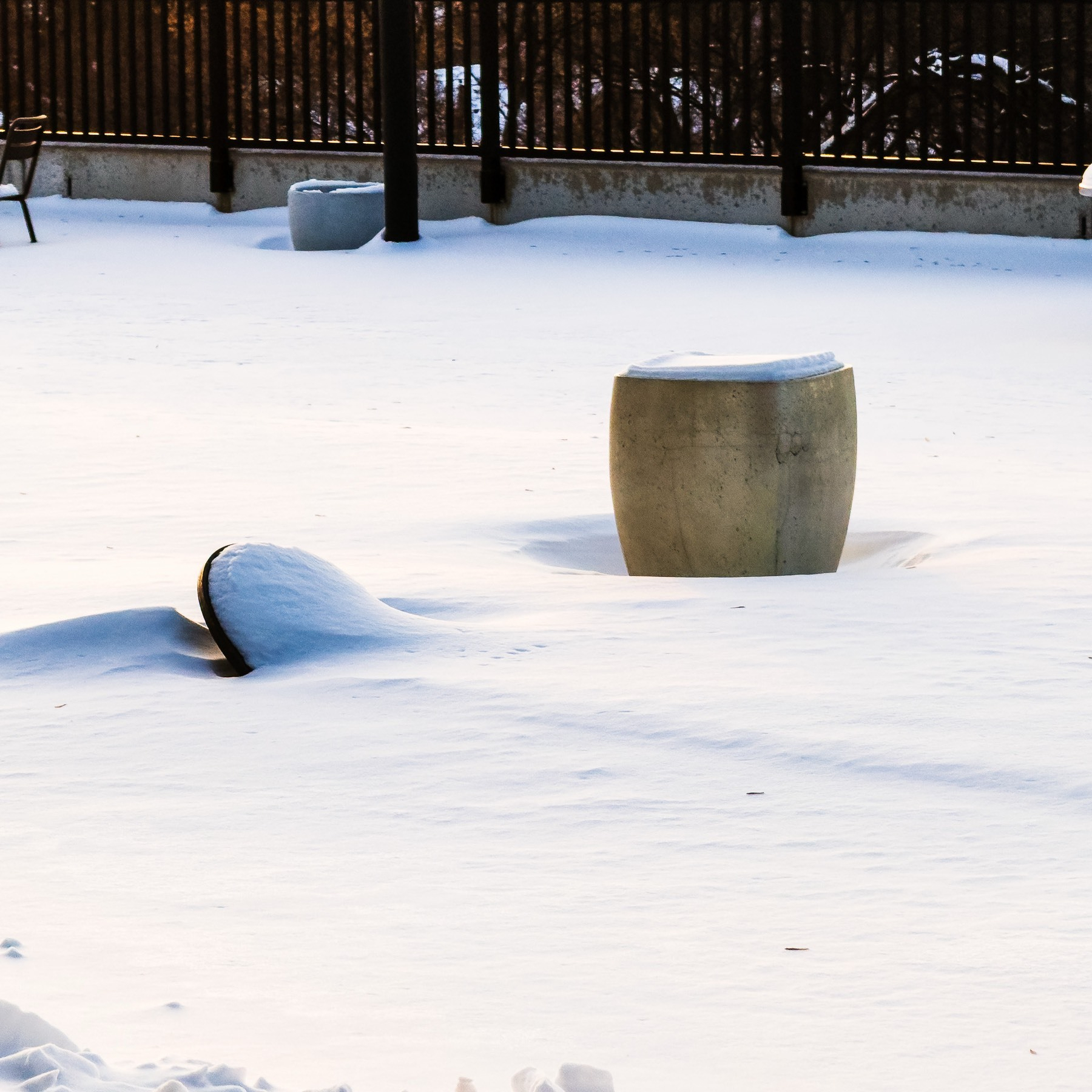 Winter scene of a snow-covered patio or garden area with large ceramic planters, one upright barrel-shaped pot with snow accumulated on top and another tipped on its side nearly buried in deep snow, with a dark metal railing and deck structure visible in the background under warm low-angle sunlight casting soft shadows across the pristine white snow.