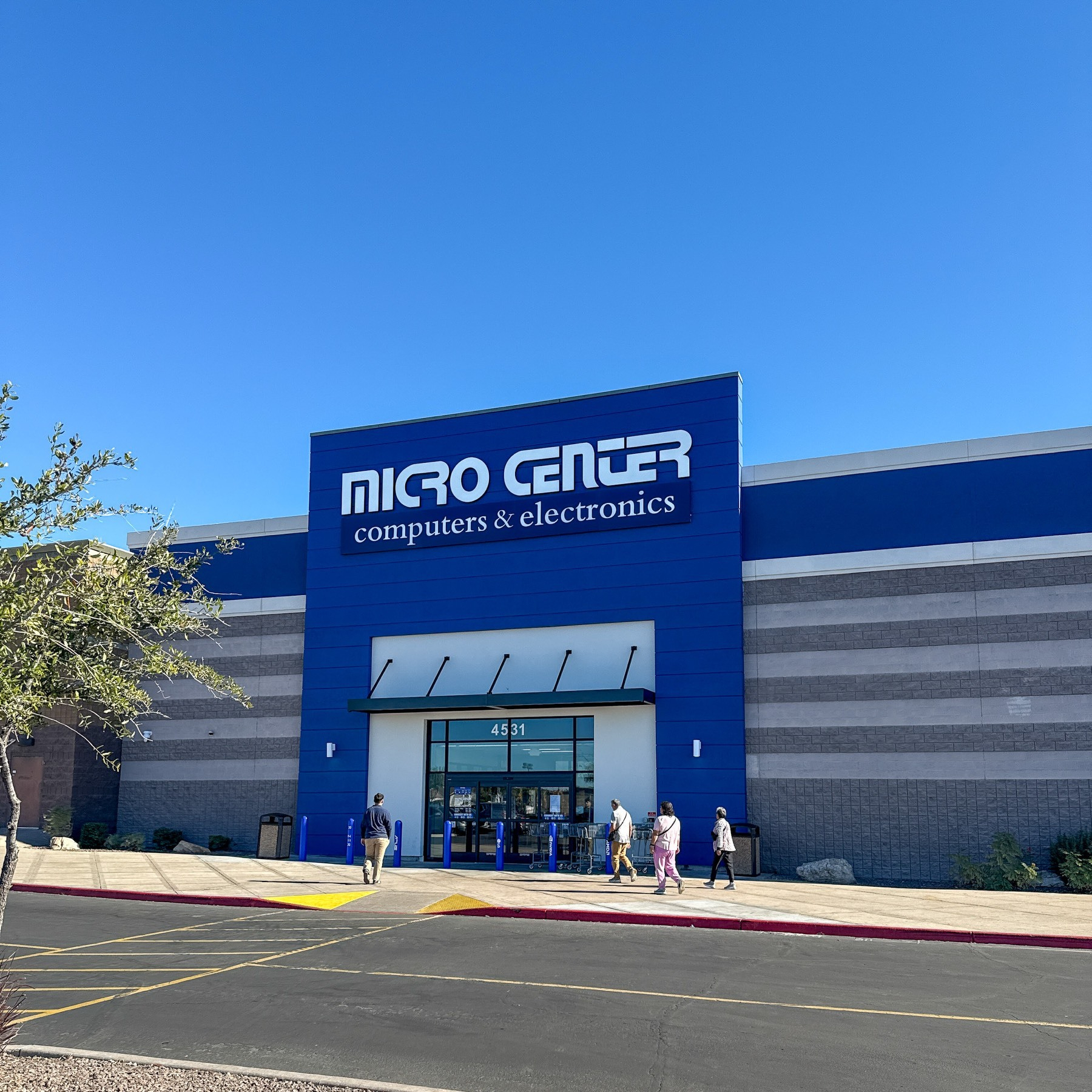 Exterior photograph of a Micro Center retail store on a clear sunny day. The building features a prominent blue facade with white signage reading MICRO CENTER and computers & electronics below. The address 4531 is displayed above the glass entrance doors. Several customers are walking near the entrance. The building has grey horizontal striped panels on either side of the blue central section, with a small tree visible on the left and a parking lot in the foreground.