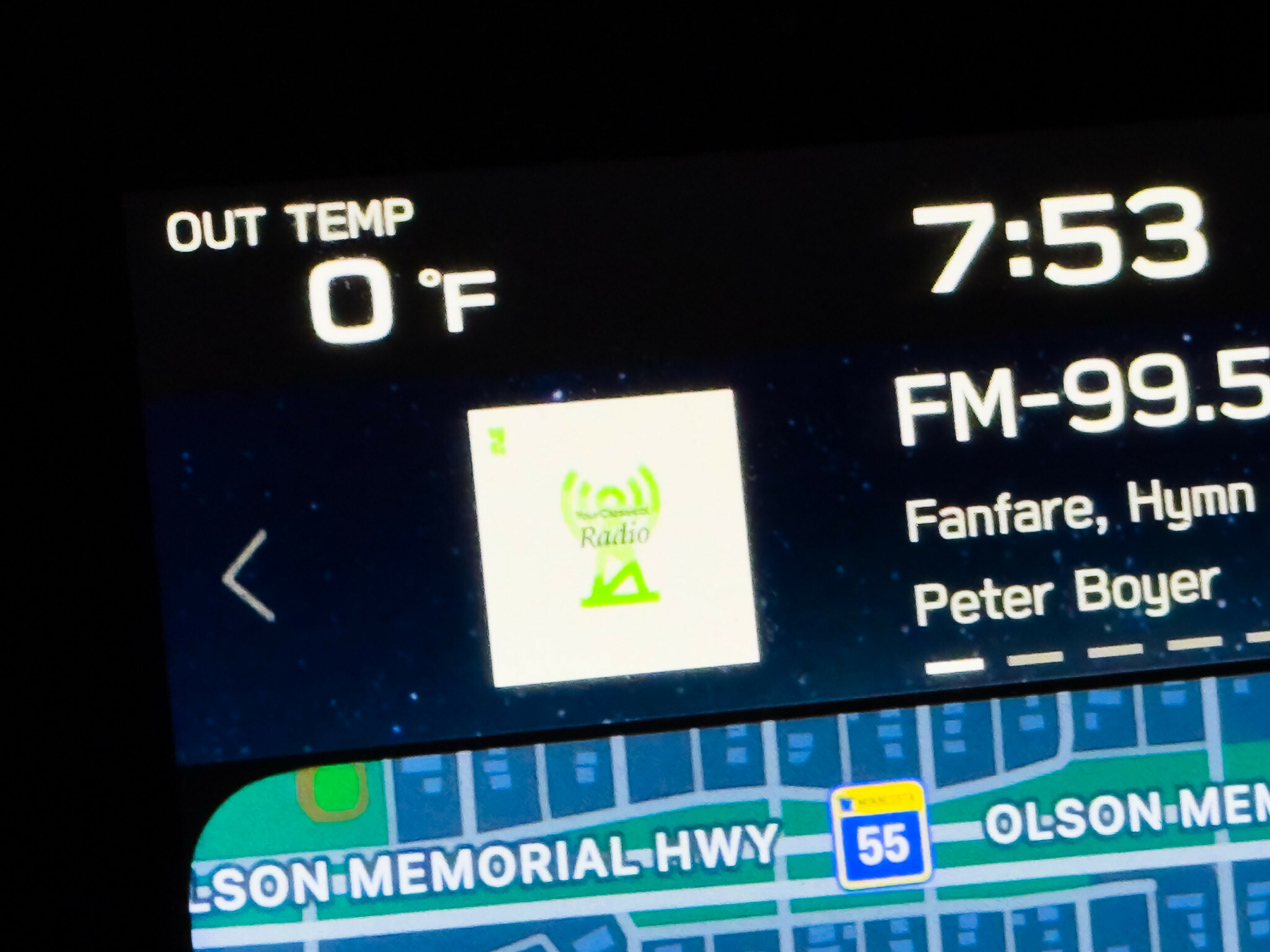 Car infotainment display showing OUT TEMP 0 °F on the left, time 7:53 in the upper right, FM-99.5 radio tuned to a classical station with a green Radio logo, displaying \"Fanfare, Hymn\" by Peter Boyer, with a navigation map at the bottom showing Olson Memorial Hwy and Route 55.