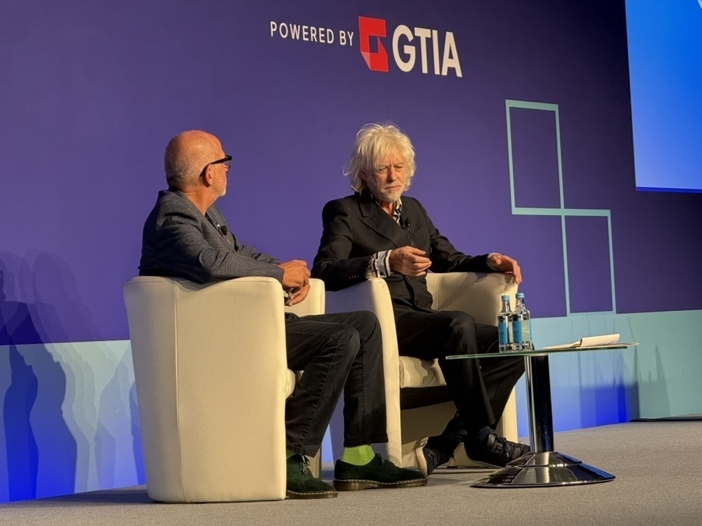 Auto-generated description: Two men seated on stage in armchairs participate in a discussion, with a blue backdrop displaying a logo.