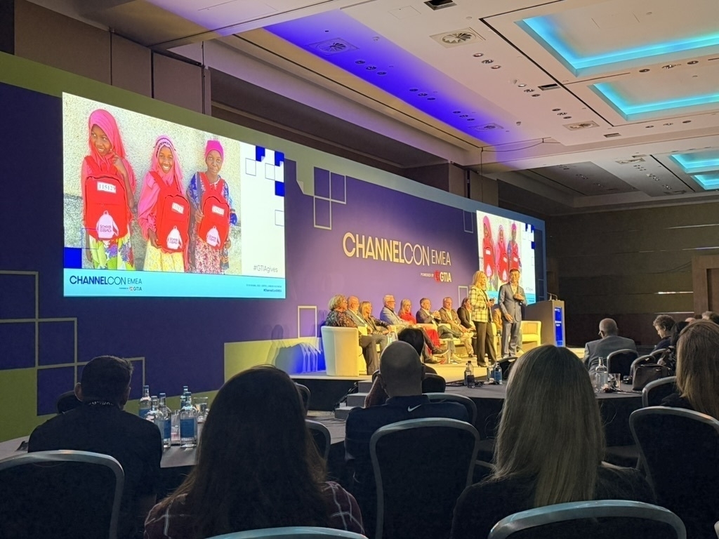 Auto-generated description: A conference or event is taking place in a large room with an audience and multiple speakers on stage, featuring a large screen displaying an image of children with backpacks.