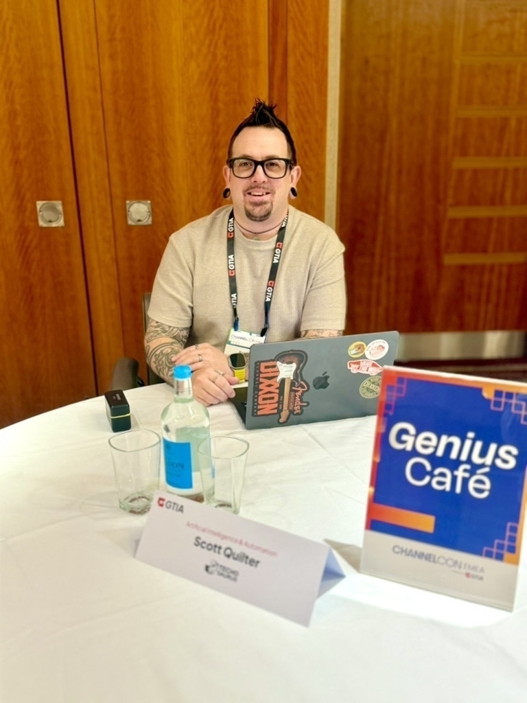 Auto-generated description: A person with tattoos is sitting at a table with a laptop, water bottle, and a sign reading Genius Café.