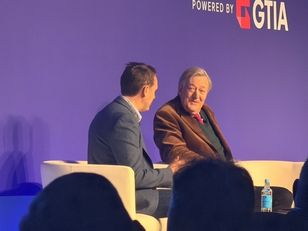 Auto-generated description: Two people are seated on stage engaged in a conversation, with a purple backdrop displaying event branding and a bottle of water on a small table.