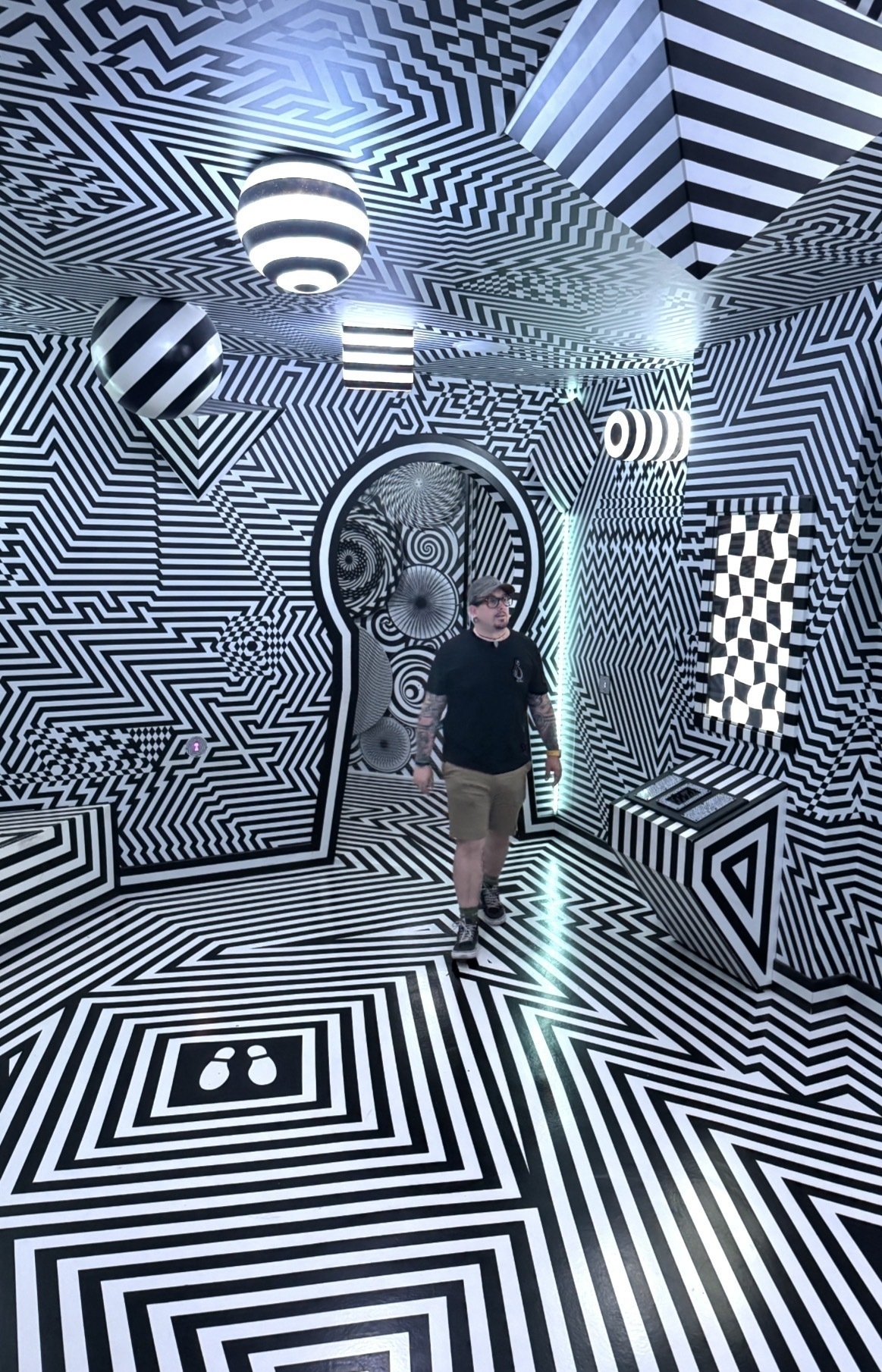 Auto-generated description: A person is walking through a room filled with dynamic black and white geometric patterns that cover the walls, ceiling, and floor.