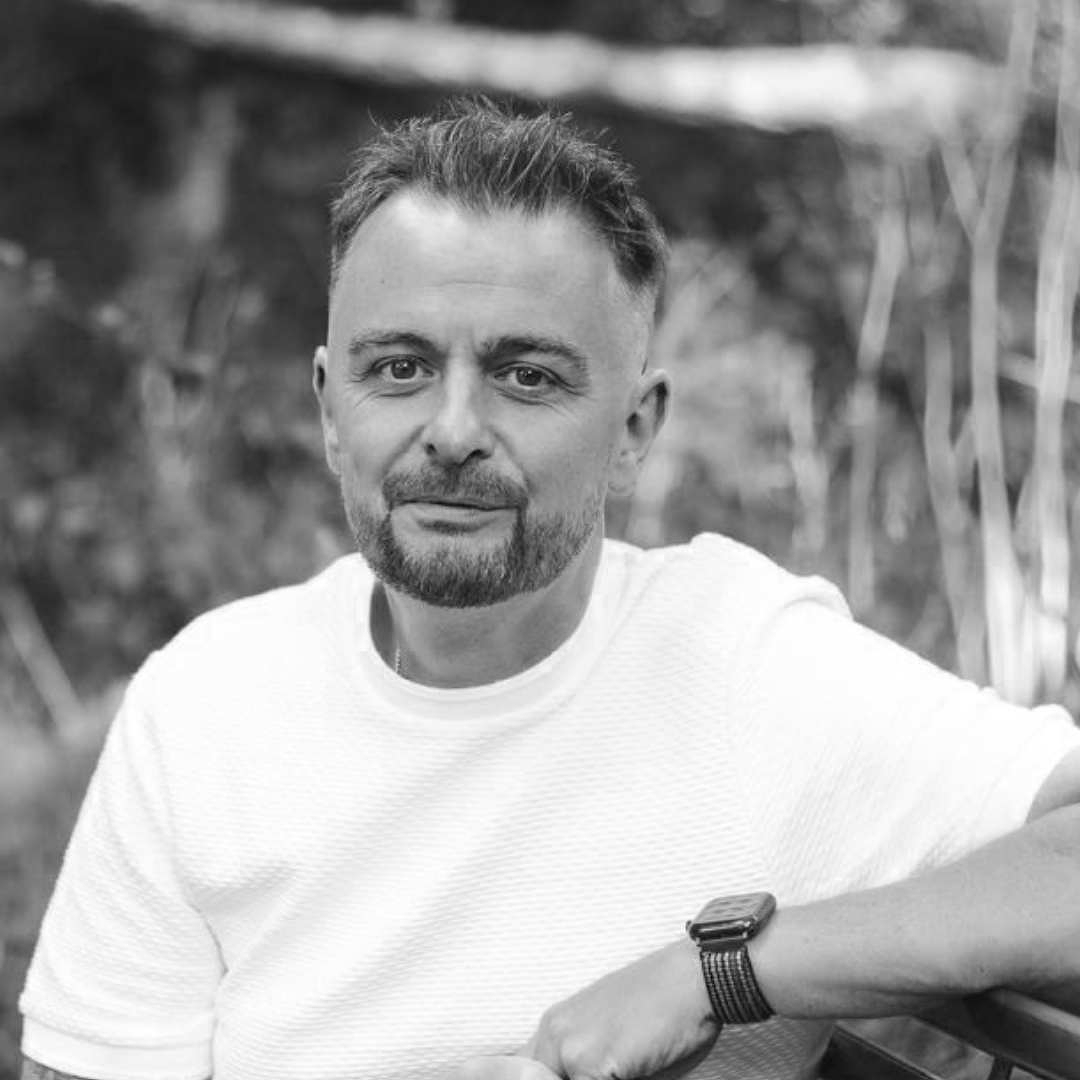 Auto-generated description: A man with short hair and a beard, wearing a white shirt, is seated outdoors, looking at the camera.