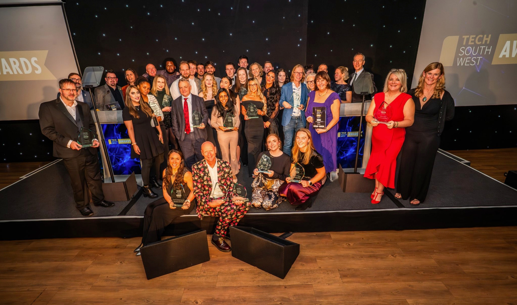 Auto-generated description: A group of people in formal attire are posing on stage with trophies at an awards event.
