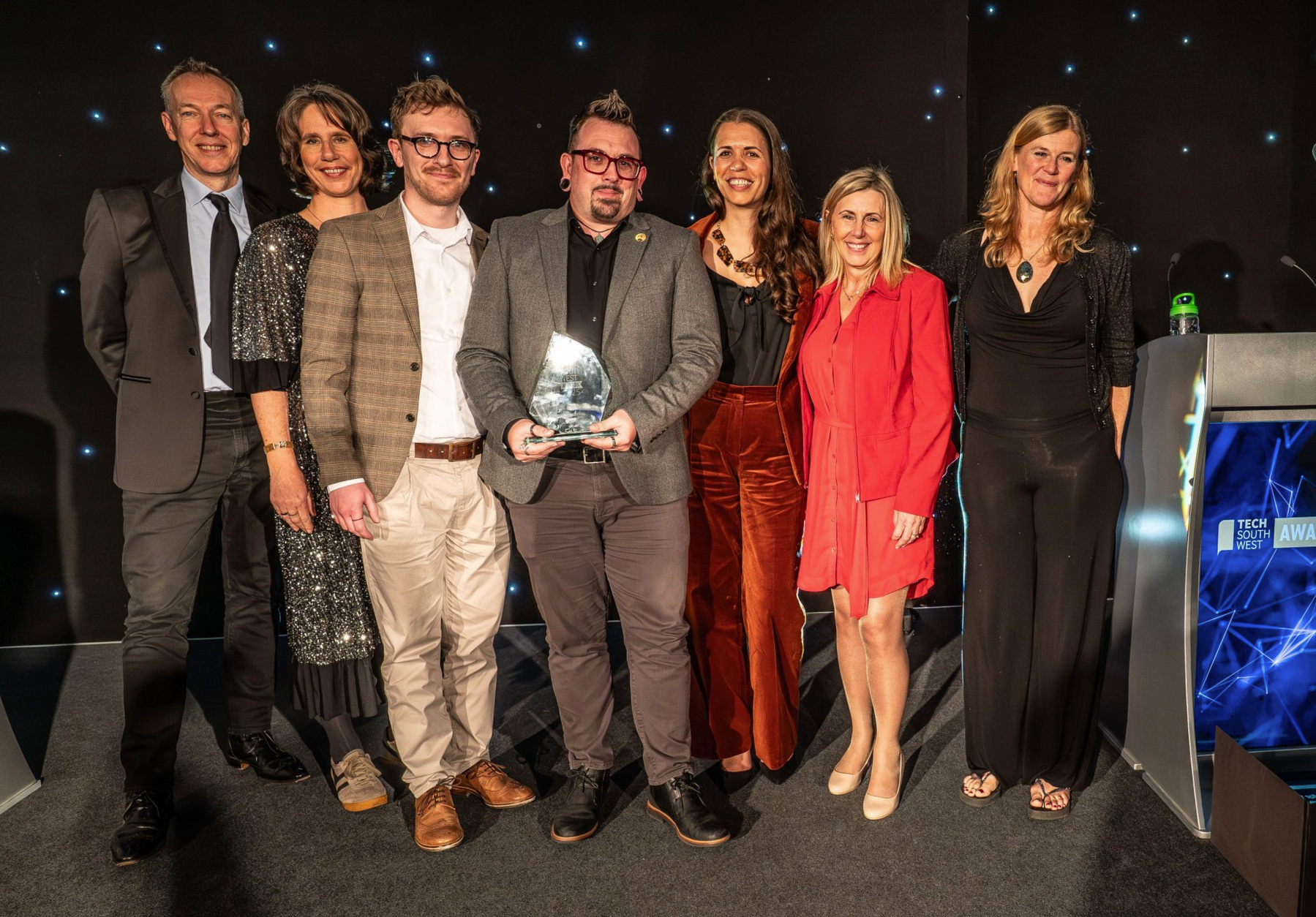 Auto-generated description: A group of people, dressed formally, stand together on a stage, and one person is holding an award.