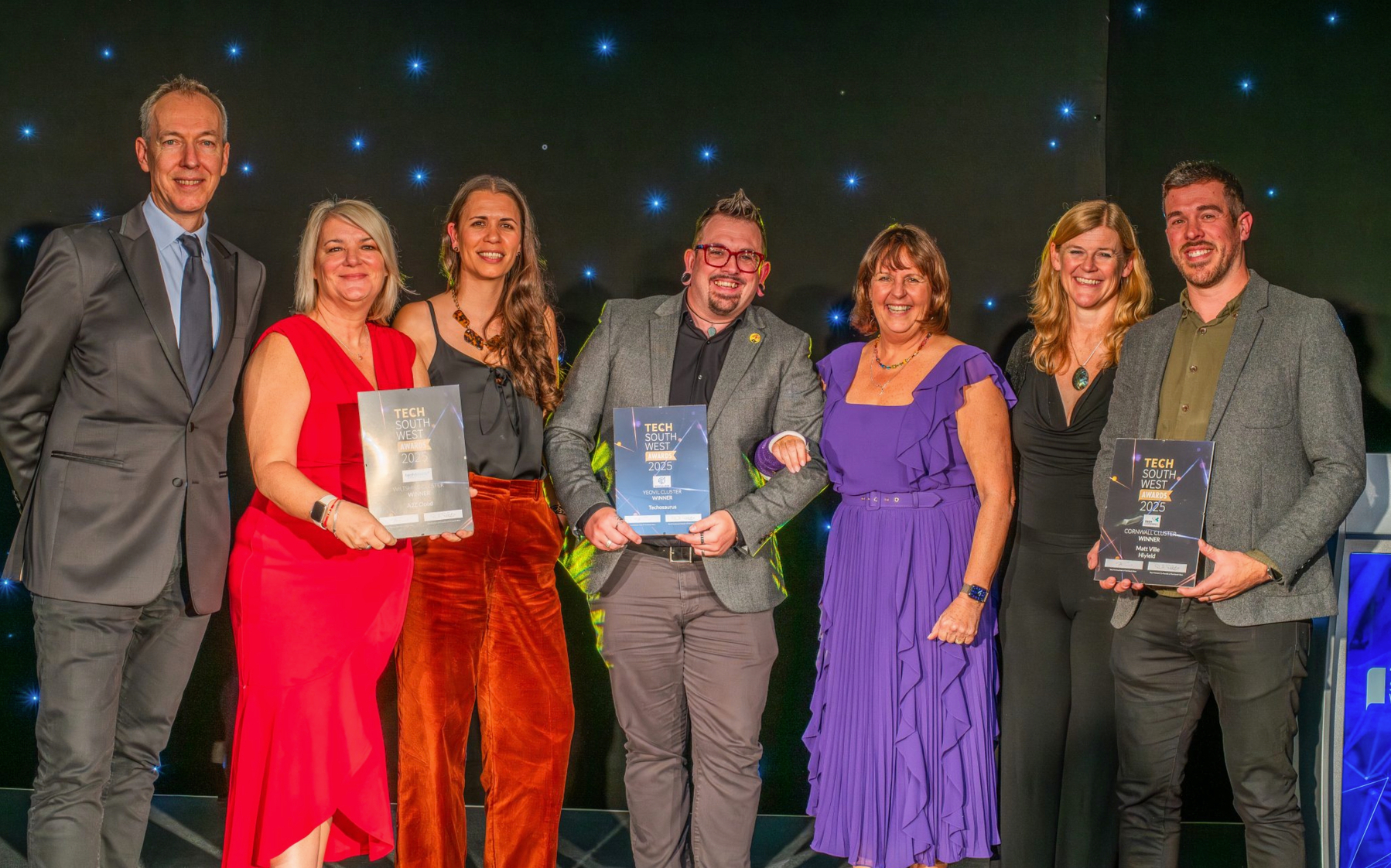 Auto-generated description: A group of seven people, some holding awards, stand together on a stage in formal attire.