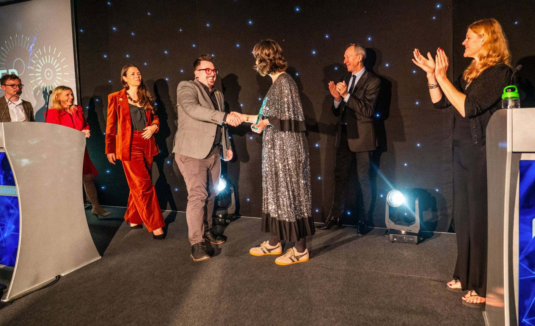 Auto-generated description: Several people on stage are engaging in a handshake and applause during an award ceremony.