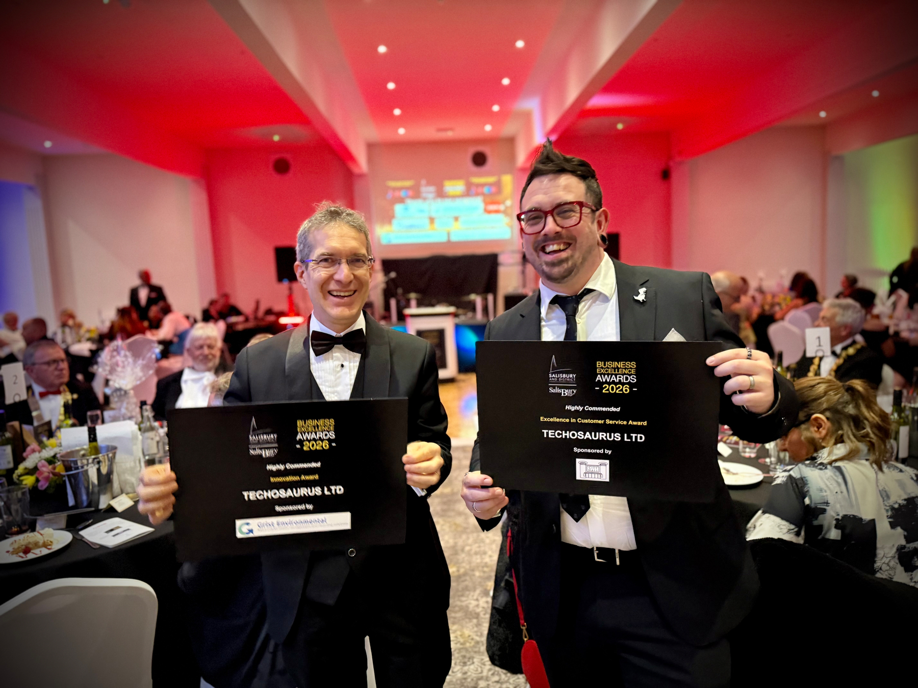 Scott Quilter and Andrea Penna of Techosaurus in formal attire hold award plaques at a festive event, surrounded by seated guests.