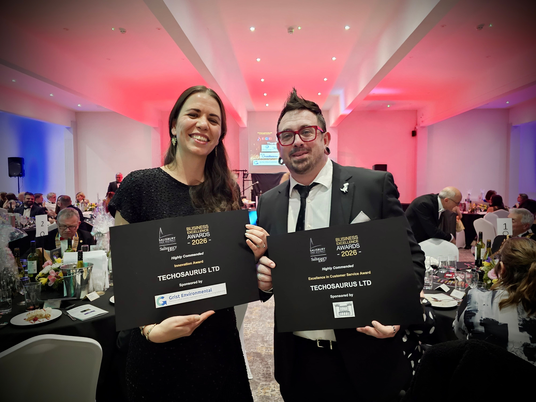 Scott Quilter and Ellie Quilter are holding award certificates in a formal event setting with other attendees and tables in the background.