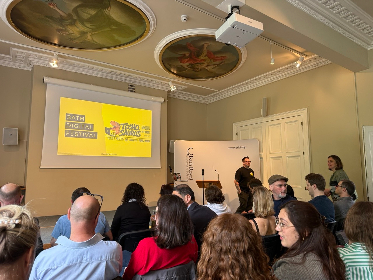 Auto-generated description: A group of people is seated in a room with ornate ceiling decorations, listening to a presentation about Bath Digital Festival and Technosaurus.