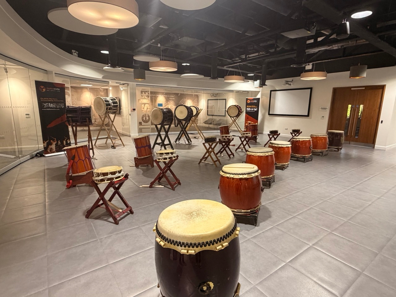 Auto-generated description: A room is set up with a variety of Taiko drums on stands and chairs arranged on a tiled floor, surrounded by display banners and lit by overhead lights.