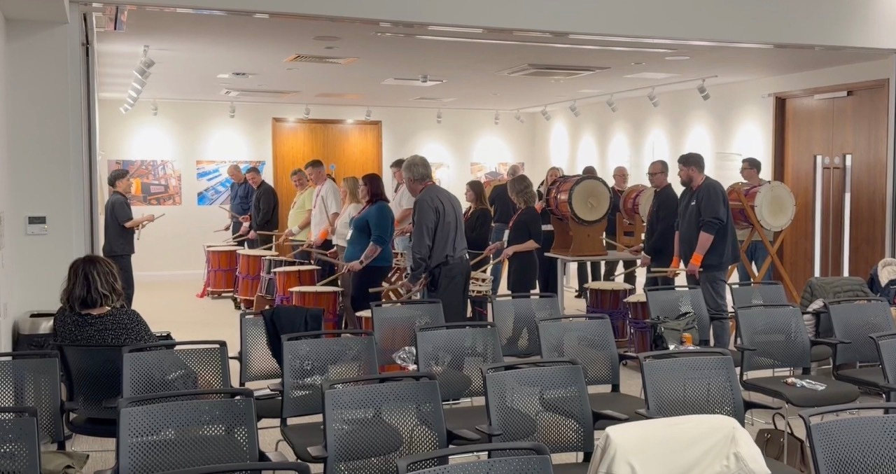 Auto-generated description: A group of people is participating in a drumming session in a room with empty chairs in the foreground.