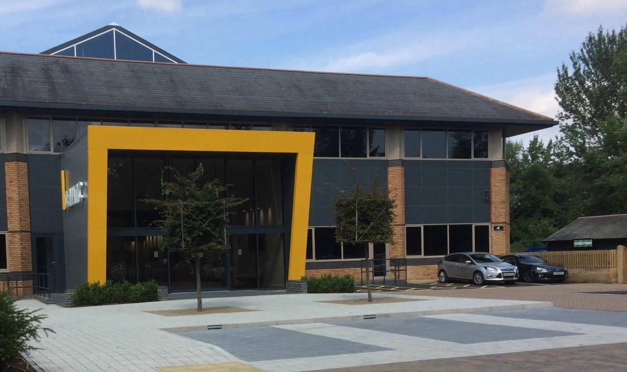 Auto-generated description: A modern commercial building with a distinctive yellow-framed entrance is surrounded by a paved plaza and parked cars.