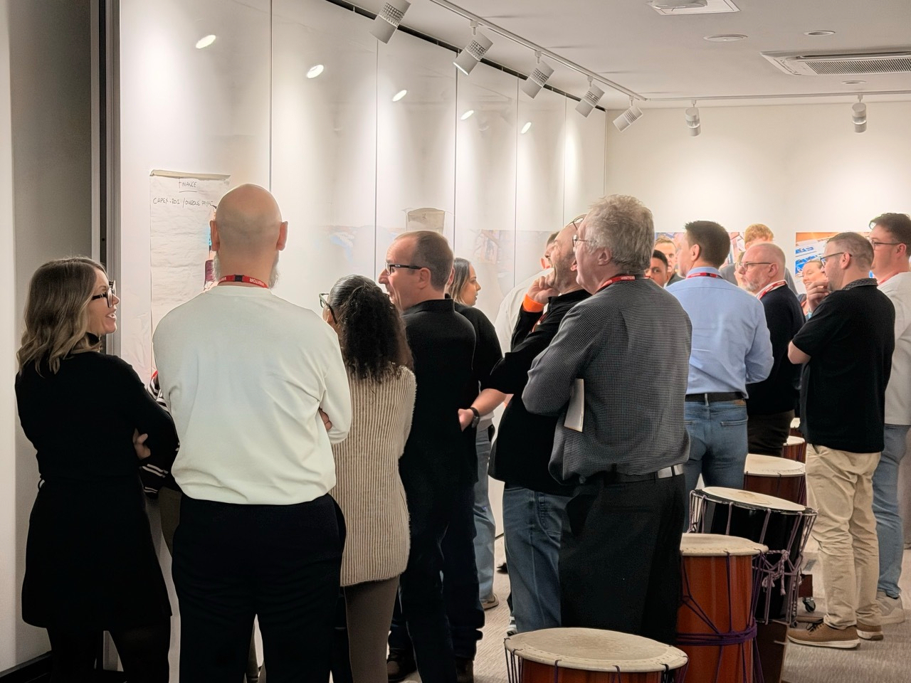 Auto-generated description: A group of people are gathered in a room, attentively looking at information displayed on boards while several drums are placed nearby.