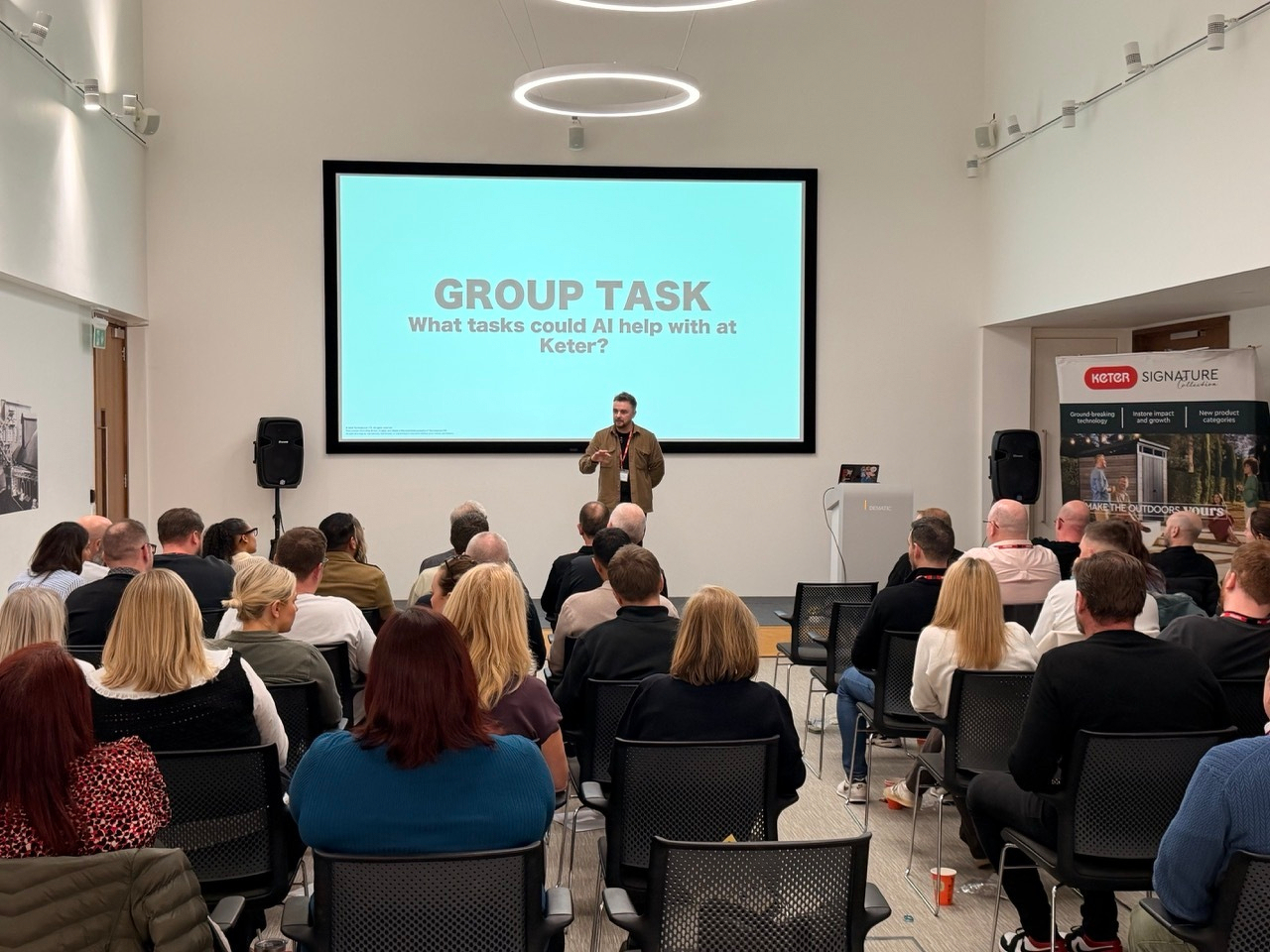 Auto-generated description: A speaker is addressing an audience in a conference room, discussing how AI could assist at Keter.