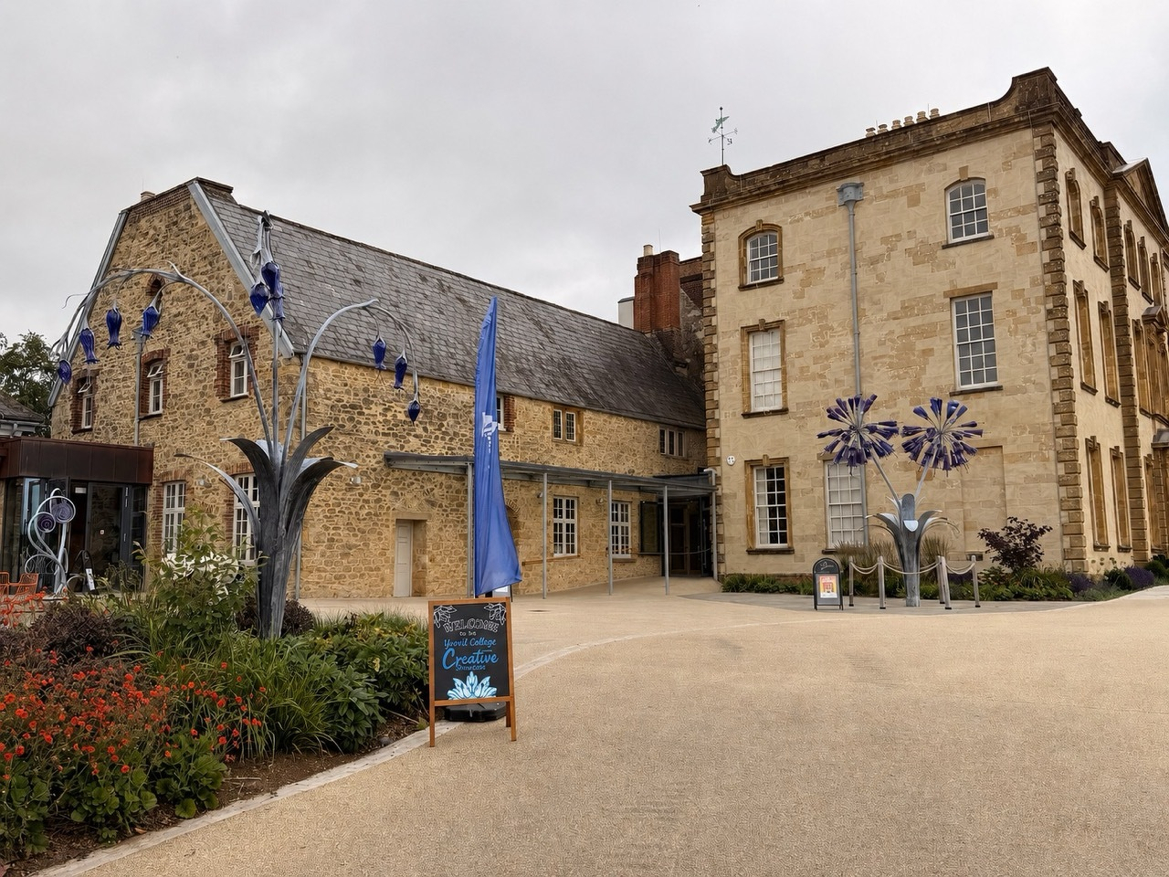 Auto-generated description: A courtyard with sculptures resembling flowers is located between two buildings, one made of stone and the other made of brick.