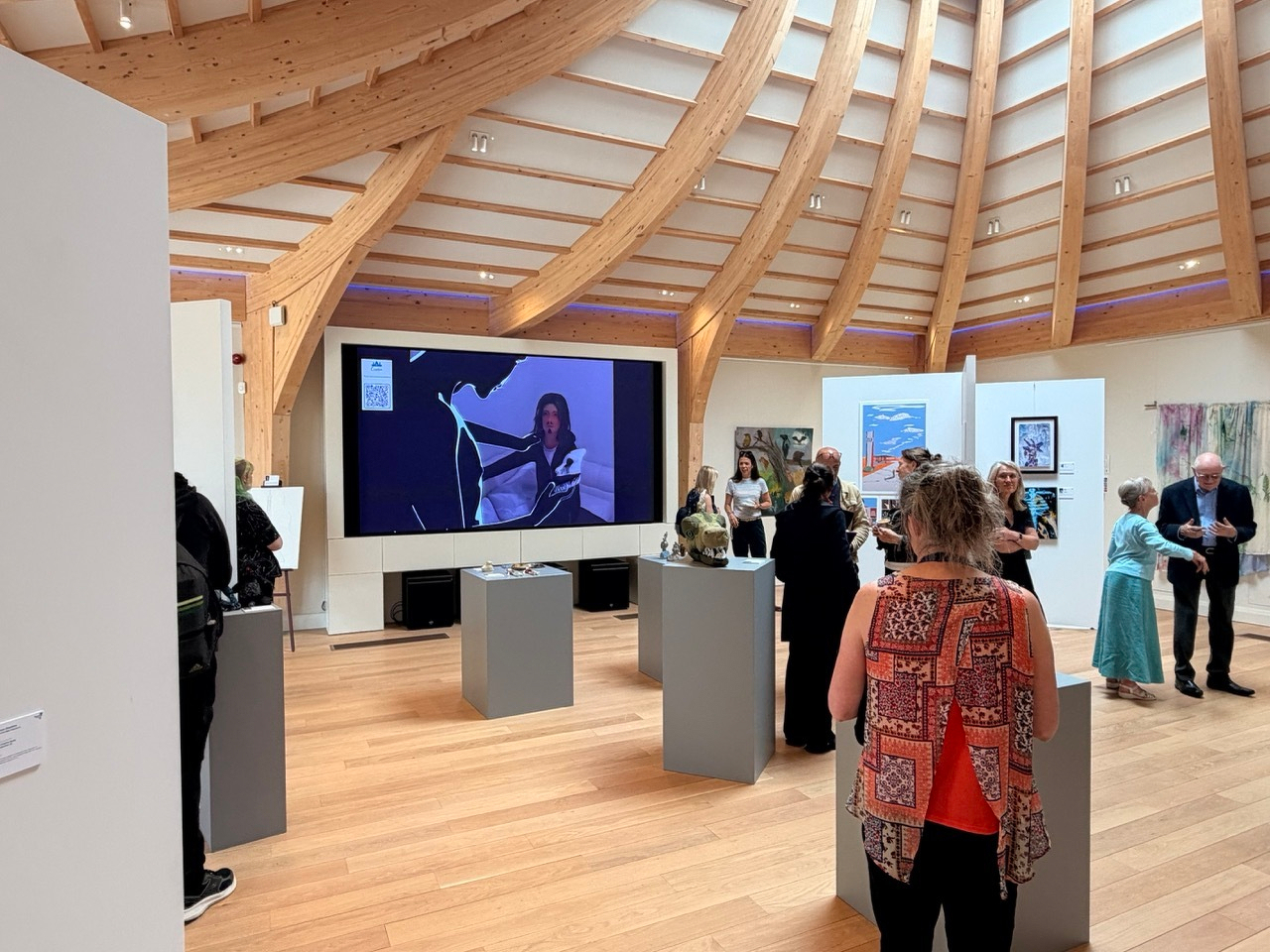 Auto-generated description: A group of people are gathered in an art gallery with wooden beams on the ceiling, viewing artwork displayed on walls and pedestals, while a large screen shows a woman speaking or presenting.