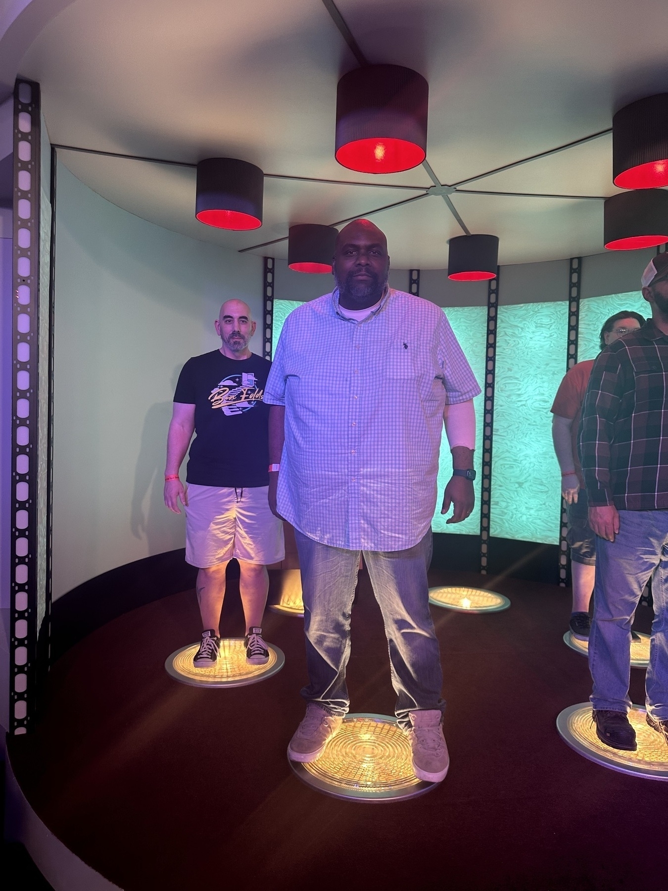 Three people are standing on lit circular platforms in a room with overhead cylindrical lights.