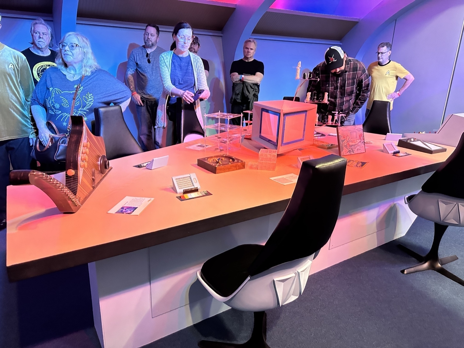 A group of people are standing around a futuristic-looking desk, which is decorated with various objects and surrounded by black swivel chairs.