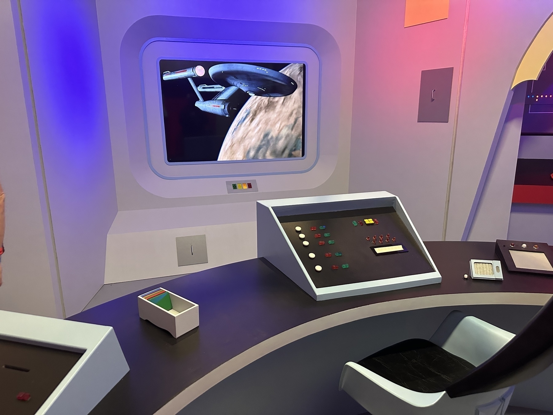 A futuristic control panel with buttons and screens is set in a room displaying a spaceship image on the wall.