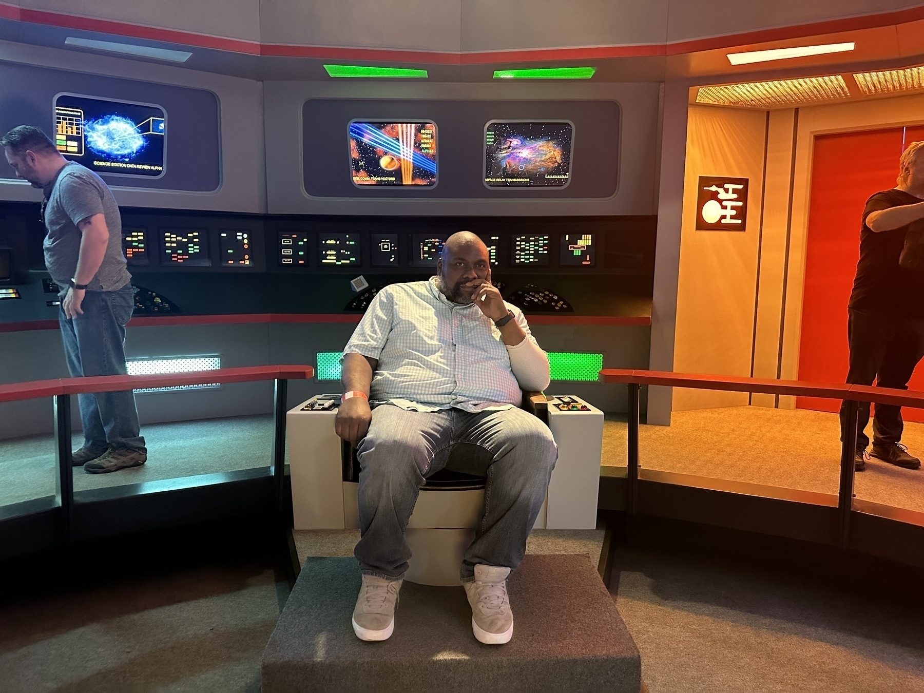 A person is sitting in a futuristic-looking chair surrounded by control panels and display screens, with two other people in the background.