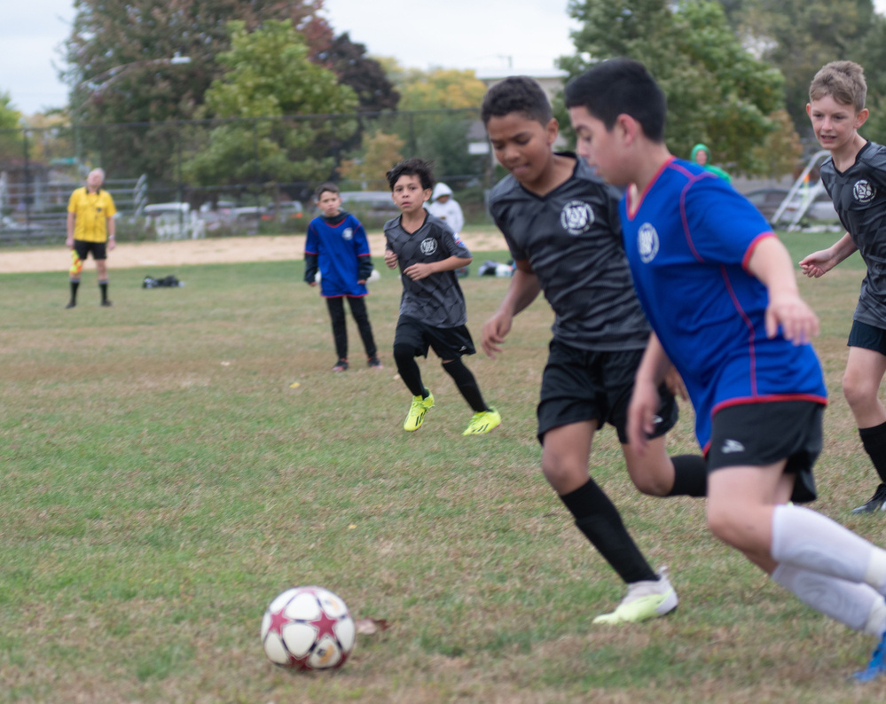 A youth soccer game is underway on a grassy field, with players in black and blue uniforms chasing the ball.