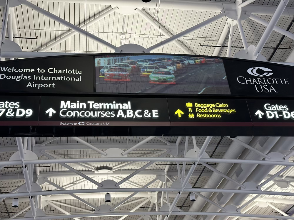 Sign, hanging from ceiling, for Charlotte International Douglas Airport, with picture of race cars in the middle and gate signs underneath.