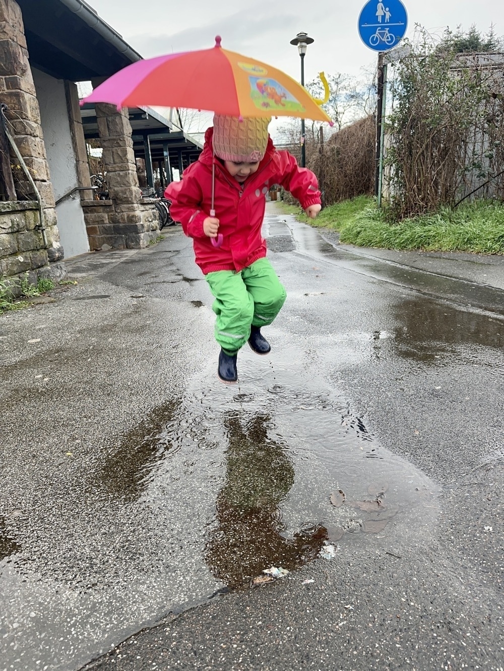 Little girl, holding an umbrella, and jumping in the air over a puddle on the sidewalk.