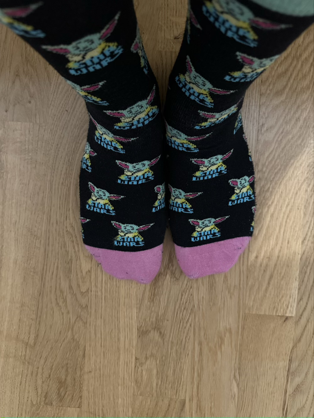 Picture of feet wearing black socks with neon pink on the toes and covered with baby Yoda designs. 