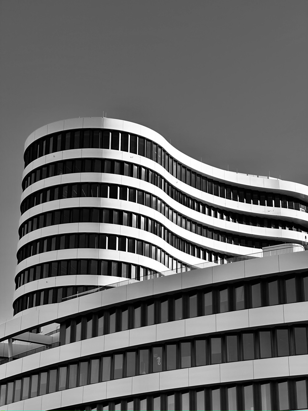 Black and white photo of a tall office building, with unique curved architecture, behind a shorter office building, with similar curved architecture. 