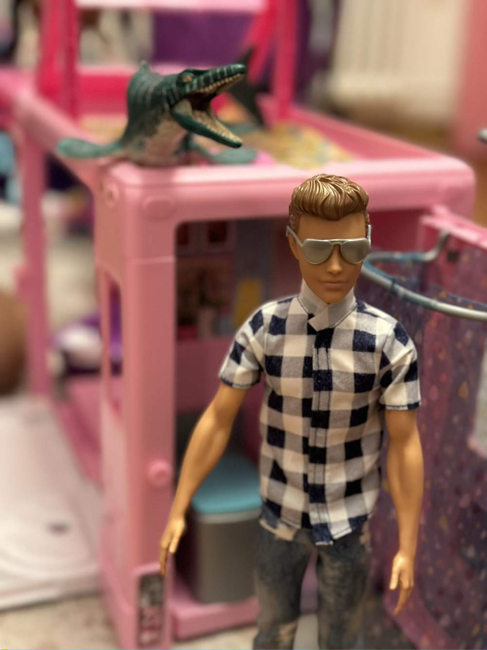 Ken doll standing in front of camper with a Mosasaur dinosaur laying on top of the camper behind him.